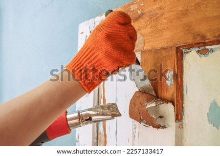The process of cleaning an old wooden door from paint layers. Removal of the coating with a hair dryer and a spatula. Worker hands in orange gloves with a tool. Household renovation. Copy space