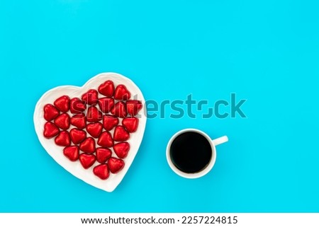 Heart shaped plate with heart shaped red foil chocolates and coffee cup on a blue background, flat lay, copy space.