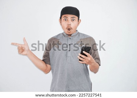 shocked young asian muslim man holding mobile phone finger pointing side wearing muslim clothes isolated on white background
