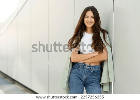 Portrait of attractive smiling woman with beautiful long hair wearing trendy casual clothing looking at camera on street, copy space. Happy fashion model posing for pictures outdoors. Natural beauty