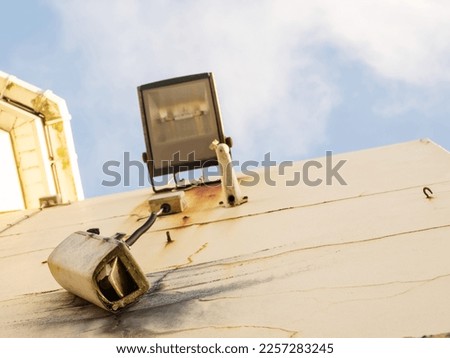 Damaged CCTV camera hanging on a cable on yellow wall, old flood light above the camera. Front element is broken and there is sign of explosion. Blue cloudy sky. War or high criminal unsafe area.
