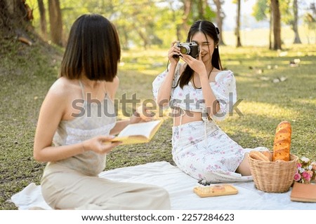 Two attractive young Asian women enjoying afternoon picnic in the park together, taking a photo from retro camera, reading a book.