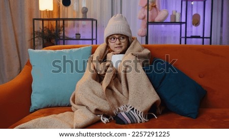 Sick toddler girl wear hat wrapped in plaid sit alone shivering from cold on sofa drinking hot cacao in unheated apartment without heating due debt. Unhealthy kid feeling discomfort try to warming up Royalty-Free Stock Photo #2257291225