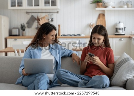 Young smiling mother talking to teen daughter child about being online, modern family using digital devices while resting on sofa at home. Mom chatting speaking with kid about internet safety Royalty-Free Stock Photo #2257299543