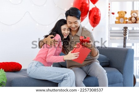 Millennial Asian young romantic lover couple male boyfriend sitting on cozy sofa giving present wrapped gift box surprise female girlfriend celebrating valentine day festival in decorated living room