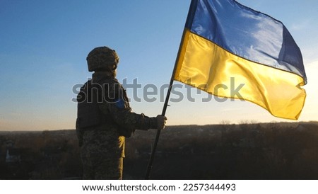 Soldier of ukrainian army holding waving flag of Ukraine against background sunset. Man in military uniform and helmet lifted up flag. Victory against russian aggression. Invasion resistance concept.