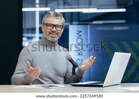Successful mature financier business coach teacher working inside office with laptop and professional microphone, senior man recording podcast and audiobook mentor looking at camera and smiling.