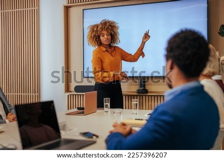 A female African American corporate manager is discussing the strategy of their business plan by pointing to a presentation on a big screen on the wall.