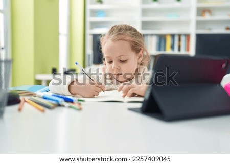 Adorable blonde girl student using touchpad writing on notebook at classroom