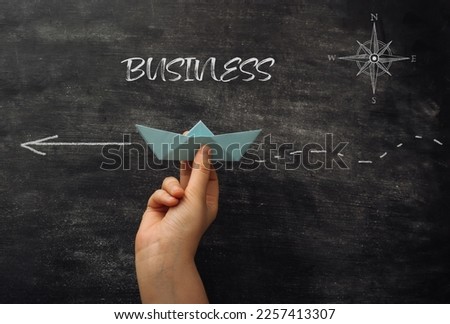 Person hand holds blue paper ship near black chalkboard with phrase Business. Right way of establishing business. Finding true and safe path Royalty-Free Stock Photo #2257413307