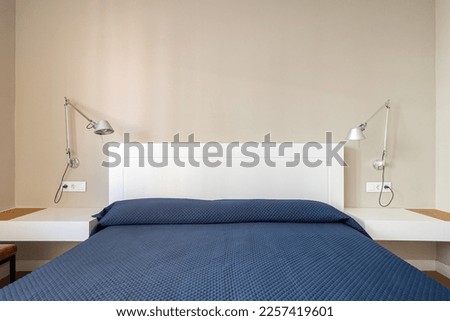 Double bed with white headboard covered with a blue bedspread. On either side are bedside tables with white tops and wall lamps with metal shades for nighttime reading. Royalty-Free Stock Photo #2257419601