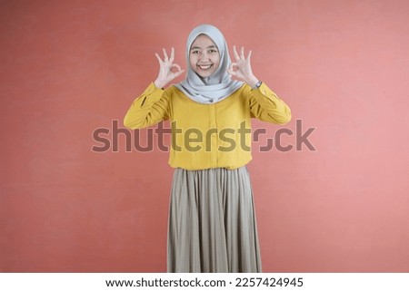 Beautiful Asian woman in yellow shirt and hijab showing good gesture showing approval symbol on brown background