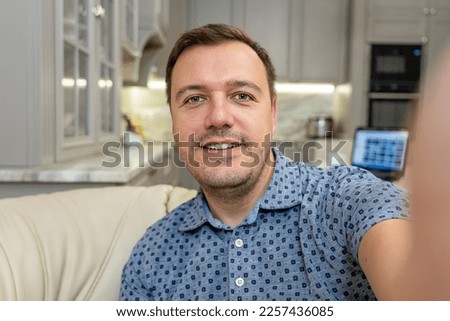 Image of millennial man smiling and taking selfie photo while sitting on sofa at home. Portrait of happy man in blue shirt taking selfie or having video call with friends showing new flat, webcam view