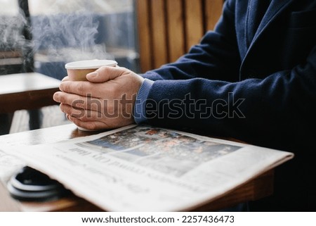 A gray-haired man in warm clothes reads a fresh newspaper and drinks hot tea or coffee on the street.