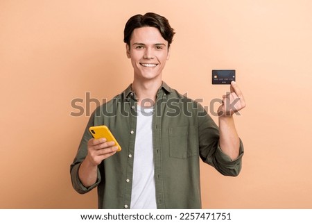 Photo of young student optimistic guy brunet hair hold his phone app for online banking hold premium plastic card isolated on beige color background