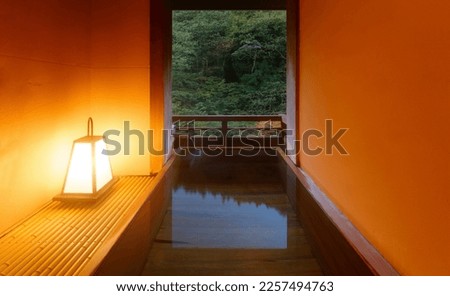 A Japanese-style open-air hot-spring bath in a traditional Onsen Ryokan, with a wooden bathtub illuminated by a lantern at blue dusk and a relaxing vibe in the natural setting of a green forest Royalty-Free Stock Photo #2257494763