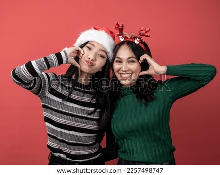 Two Asian Indonesian women in Christmas attire look cute as they make a heart gesture with their hands on their cheeks. Isolated on red background