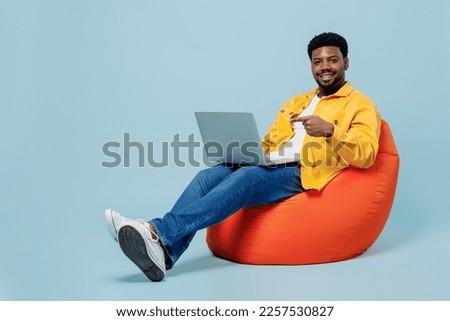 Full body happy young man of African American ethnicity wear yellow shirt sit wearing bag chair hold use work point finger on laptop pc computer isolated on plain pastel light blue background studio.