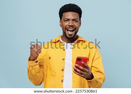 Young man of African American ethnicity 20s in yellow shirt hold in hand use mobile cell phone do winner gesture isolated on plain pastel light blue background studio portrait People lifestyle concept