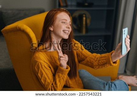 Happy young woman taking selfie picture or chatting by video call showing victory sign on mobile phone sitting in cozy yellow armchair at home. Redhead female communicating online using smartphone.