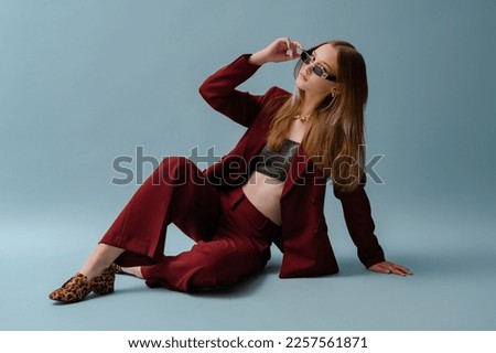 Fashionable confident woman wearing elegant marsala color suit, leather crop top, leopard print loafer shoes, posing on blue background. Full-length studio fashion portrait. Copy, empty space for text