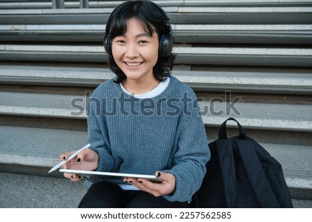 Young asian girl, student in headphones, works on remote, digital artist drawing on tablet with graphic pen, listening music in headphones and sitting on street staircase.