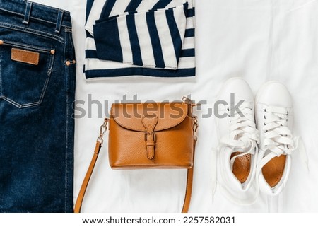 Blue jeans, t shirt, bag and white sneakers on white bed. Trendy clothes. Women's casual spring summer outfit. Fashion concept. Flat lay, top view Royalty-Free Stock Photo #2257582031