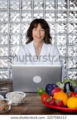 Smiling female nutritionist in medical uniform, looking at camera, surfing internet on netbook while working at desk with fresh vegetables and dried legumes. Front view, Vertical