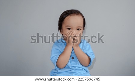 cute asian toddler wearing blue tshirt closing his mouth by hands over grey.
