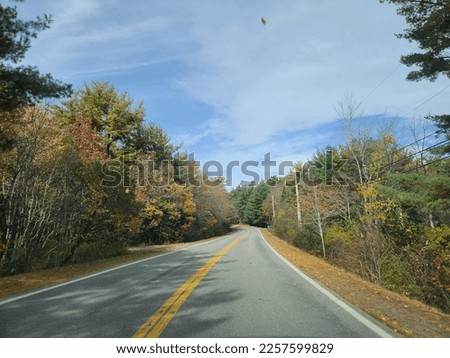 The view of a well cared for stretch of highway under a blue sky with clouds stretched over it. The empty two lane highway is surrounded by fall colored trees on either side. The leaves are bright.