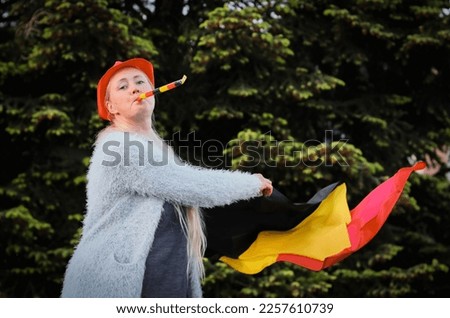 Portrait of a beautiful blonde middle-aged caucasian woman with a cap on her head waving the Belgian flag and blowing a whistle celebrates independence day and victory in a football match in the backy