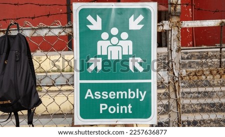 a poster that reads assemble point attached to a wire fence at a sports field.