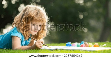 Close up face of cute child outdoors. Spring banner for website header. Artist kids. Kid draws in park laying in grass having fun on nature background.