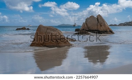 Picturesque granite rocks in the surf on a tropical island. The waves of the turquoise ocean spread along the beach. Reflection on the shiny wet sand. The yacht is far away. Clouds in the blue sky. 