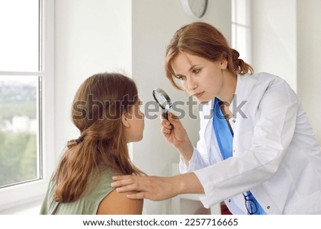 Female doctor examining child's skin. Professional dermatologist in white coat uses magnifying glass to investigate and diagnose some growth on face of teenage girl. Dermatology, skin health concept Royalty-Free Stock Photo #2257716665