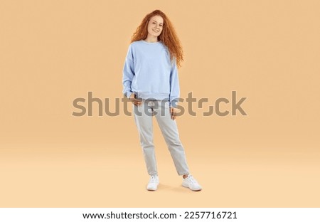 Portrait of beautiful and cute teenage girl in casual clothes on beige background in studio. Caucasian girl with red wavy hair, wearing sweatshirt and jeans, is smiling at camera. Full length.