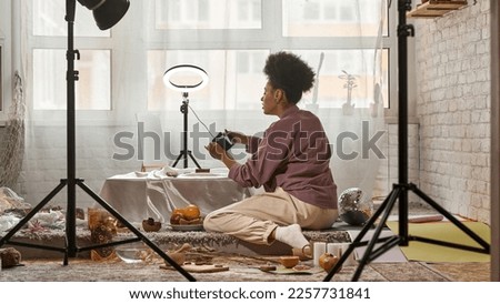 Distance view of black female photographer taking photo of seashells on digital camera at home studio. Young girl creating content for photostocks, commerce, social networks and advertising. Freelance