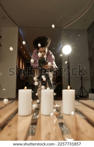 Black female photographer taking photo of falling pearls on wooden table with burning candles on digital camera on tripod at studio. Content for photostocks, commerce, social networks and advertising