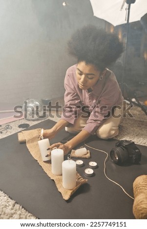 Black female photographer ligting candles for taking photo with deodorant and sack at home studio. Creating beauty and body care content for photostocks, commerce, social networks and advertising