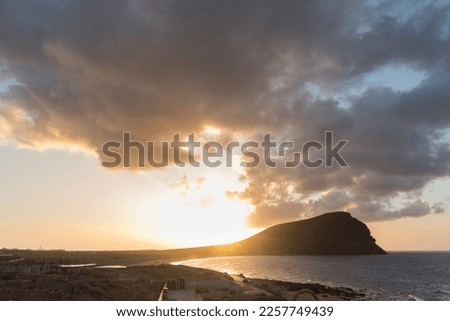 Sunset on the beach, golden sunbeams breaking through the gray storm clouds, with Red Mountain and ocen in the background. La Tejita, Tenerife, Canary Islands. Spain