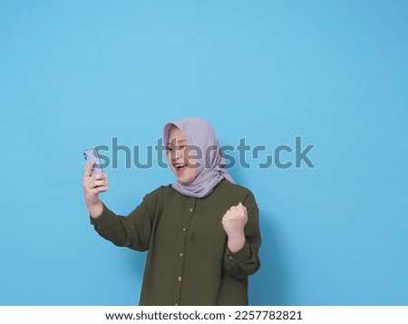 Potrait of Asian happy muslim woman wearing green long sleeve shirt and grey hijab checking the smartphone and fist her hand isolated on blue background