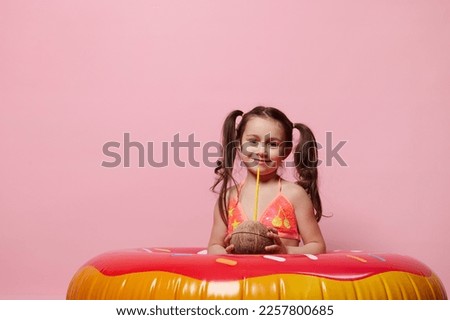 Adorable Caucasian little child girl with inflatable donut swimming ring, holding a coconut cocktail, cutely smiling looking at camera, isolated over pink color background. Summer and beach concept