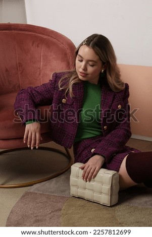 Fashionable girl sitting with a beige squared bag on floor near chair. fashionable beige squared bag closeup. Hands holding bag. Woman vilet outfit, jacket and skirt