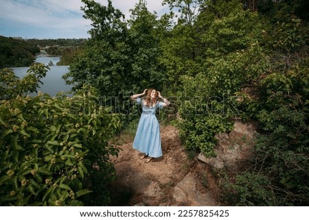 A young girl with curly blond hair, in a blue dress, laughs and touches her hair with her hands, on a rock against the backdrop of trees and a picturesque river.