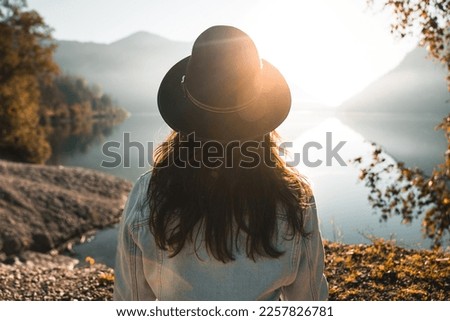 Back view of fashionable young woman with long hair, stylish denim jacket and sleek black hat looking at view against lake reflections and sun beam. Calm water, serene atmosphere, breathtaking scenery Royalty-Free Stock Photo #2257826781
