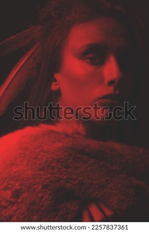 Beautiful woman portrait with dreadlocks and feathers attached to her hair. Model with make-up looking at camera and wearing animal fur coat. Toned image with red and black color
