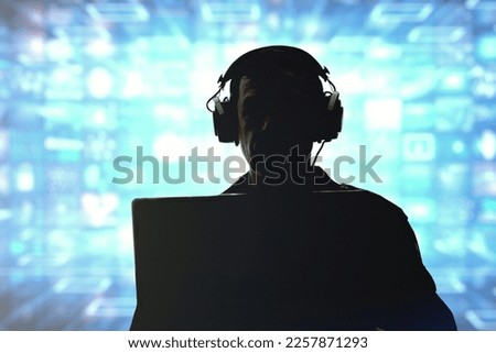 Silhouette of a military man in headphones at a laptop against the background of a video wall of glowing screens, contour lighting. Concept: information gathering, surveillance and control. Royalty-Free Stock Photo #2257871293