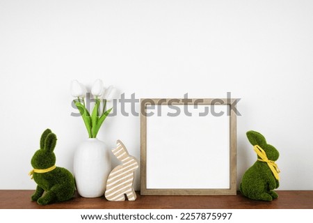 Mock up wood frame with Easter decor on a wood shelf. Moss and knit bunnies and tulip flowers. Square frame against a white wall. Copy space.
