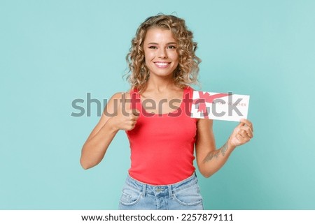 Smiling young blonde woman 20s wearing pink basic casual tank top standing hold in hand gift certificate showing thumb up looking camera isolated on blue turquoise colour background studio portrait