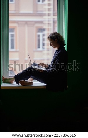 Profile shot of female in business suit looking at laptop during working process sitting on the window in co-working open office space on the dark background. High quality photo